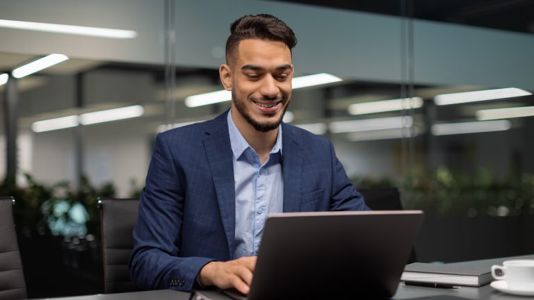 Businessman in a modern office smiling while reading tax updates on his laptop.